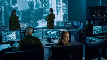  A Global Security Operations Center where various operators look at computer screens and a large video wall simultaneously. 