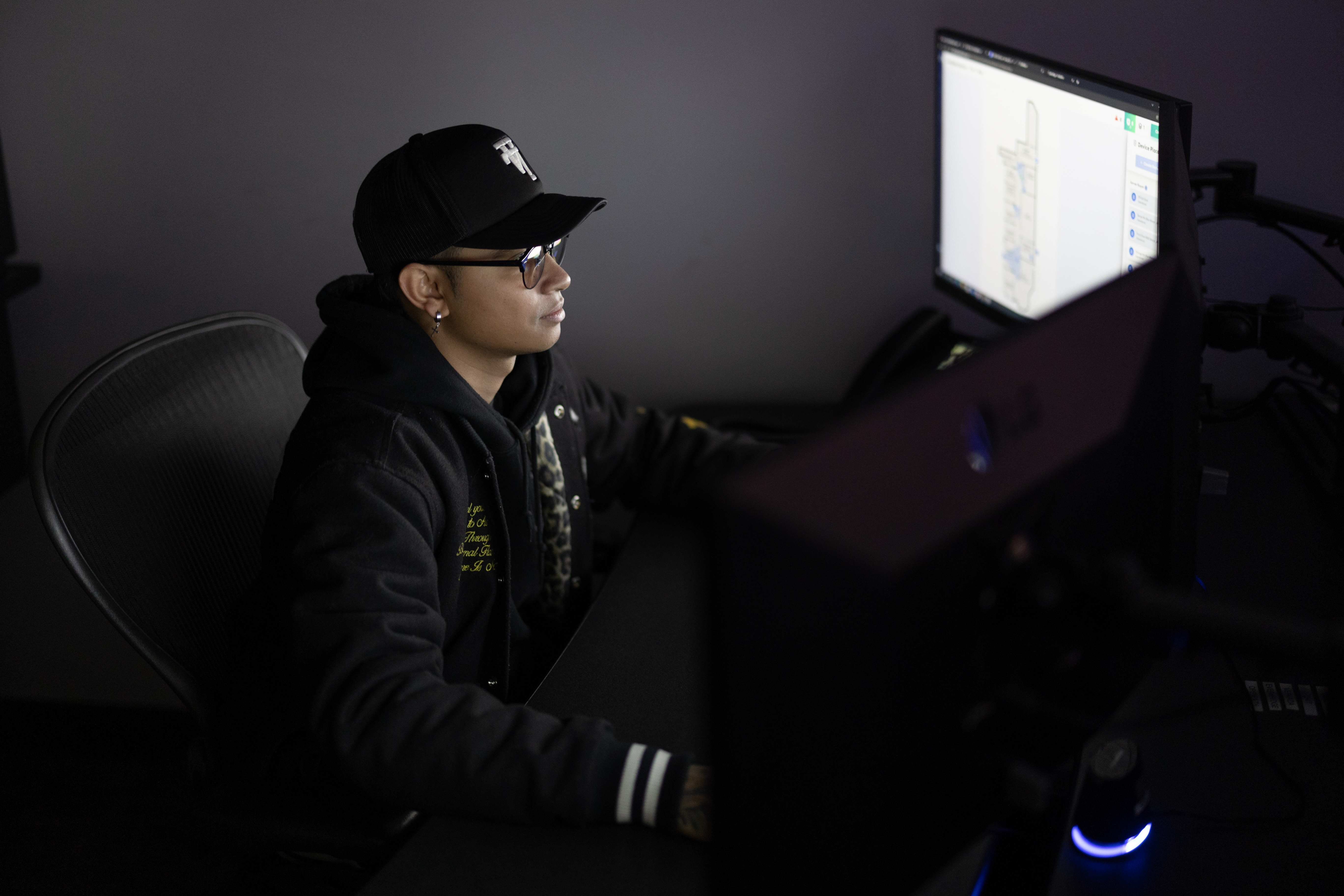 A security operator views a screen in a dark and shadowy global security operations center, wearing glasses, a black hat and black hoodie.